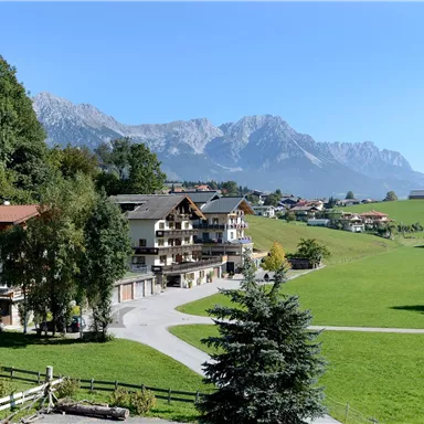 Eine malerische Landschaft mit Häusern im Vordergrund und majestätischen Bergen im Hintergrund. Die Wiese ist grün und weitläufig, unter einem strahlend blauen Himmel.