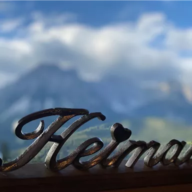 Eine schöne Landschaft mit Bergen im Hintergrund. Im Vordergrund steht das Wort "Heimat" in geschwungener Schrift.