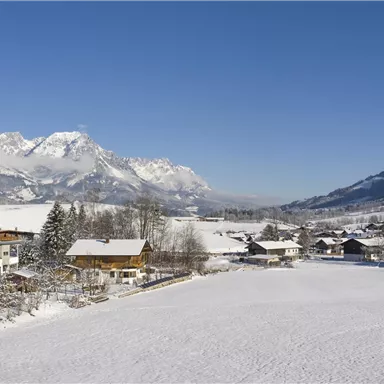 Eine schneebedeckte Landschaft mit malerischen Häusern und Bergen im Hintergrund. Der Himmel ist klar und blau.