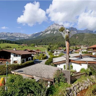 Eine malerische Alpenlandschaft mit typischen Holzhäusern und grünen Wiesen. Im Hintergrund erheben sich majestätische Berge unter einem blauen Himmel mit Wolken.