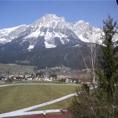 Eine malerische Berglandschaft mit schneebedeckten Gipfeln und klarem, blauem Himmel. Im Vordergrund sind grüne Wiesen und einige Gebäude zu sehen.