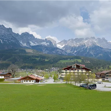A picturesque landscape with high mountains and a clear sky. In the foreground, there is a green meadow and a small settlement with houses.