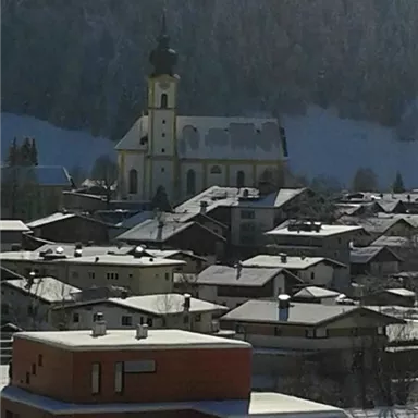 A picturesque village scene with snow-covered houses and a church in the background. The snow-covered landscape is surrounded by mountains.