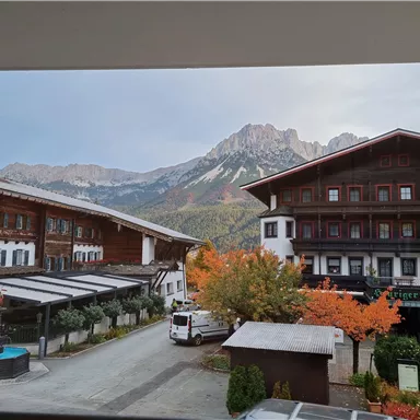 A picturesque view of the mountains with autumn trees in the foreground. The image features traditional buildings and a small road.