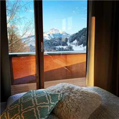 A view from a cozy room with a bed and pillows. In the background, there are snow-covered mountains and a clear blue sky.
