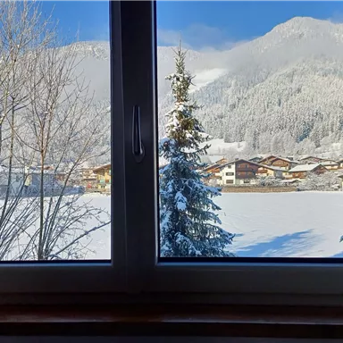 Ein Winterblick aus einem Fenster auf verschneite Berge und Bäume. Im Vordergrund sieht man ein ruhiges, schneebedecktes Landschaftsbild.