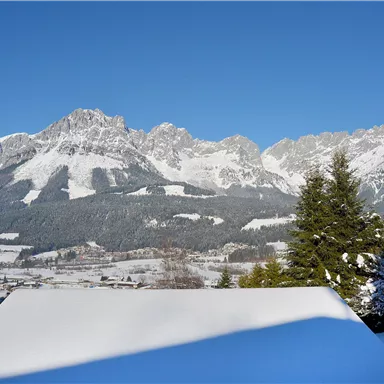 Eine schöne Winterlandschaft mit schneebedeckten Bergen und klarem blauem Himmel. Im Vordergrund sind schneebedeckte Dächer und Tannen zu sehen.