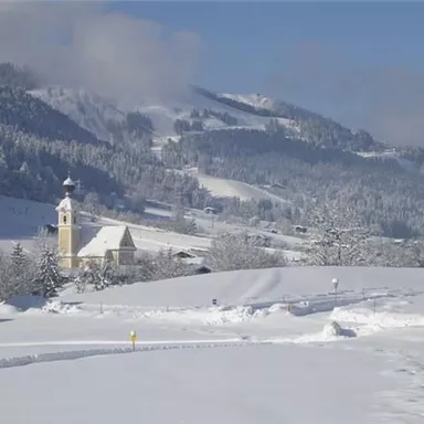 Eine verschneite Landschaft mit sanften Hügeln und einem klaren blauen Himmel. Im Vordergrund steht ein kleiner Ort mit einer Kirche.