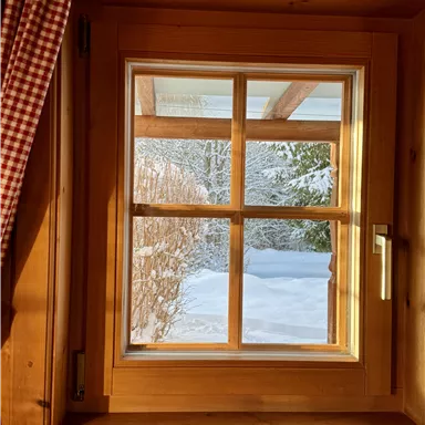 A window with a wooden frame that overlooks a snowy landscape. The gentle sun rays bathe the room in warm light.
