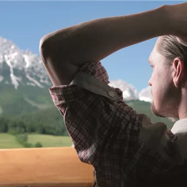 Ein Mann sitzt mit dem Rücken zur Kamera und betrachtet eine malerische Berglandschaft. Die Berge sind schneebedeckt und die Wiesen sind grün unter einem klaren blauen Himmel.