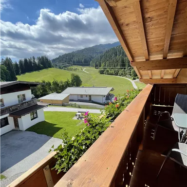 Ein schöner Blick von einem Balkon auf eine grüne Landschaft mit Hügeln und Wolken. Im Vordergrund sieht man Holzmöbel und blühende Pflanzen.