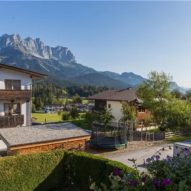 Schöne Berglandschaft mit klar blauem Himmel und grünen Wiesen. Im Vordergrund sieht man typische alpine Gebäude und einen gepflegten Garten.