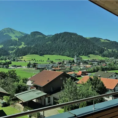 A picturesque landscape with green meadows and mountains in the background. In the foreground, there are some houses and a small church visible.