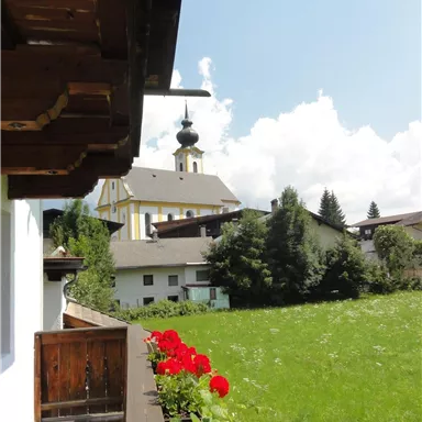 A picturesque landscape with green meadows and a small church. In the foreground, red geraniums are blooming on a balcony.