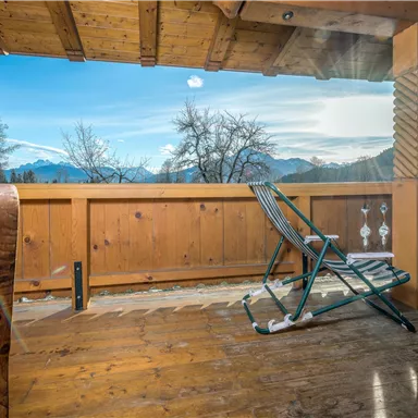 Eine gemütliche Terrasse mit Holzgeländer und Blick auf die Berge. Der Himmel ist klar und die Sonne scheint.