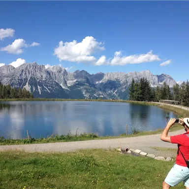Eine Person fotografiert eine malerische Berglandschaft mit einem klaren See im Vordergrund. Die Umgebung ist grün und von beeindruckenden Bergen umgeben.