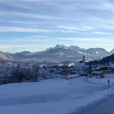 Eine winterliche Landschaft mit schneebedeckten Bergen und einer kleinen Stadt im Vordergrund. Der Himmel ist klar und blau, was die ruhige Atmosphäre unterstreicht.