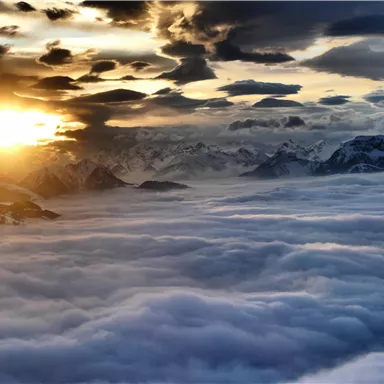 A sunrise over snow-covered mountains. The sky is filled with dramatic clouds and a sea of mist.