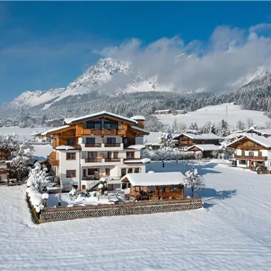 A picturesque winter landscape with snow-covered houses and mountains in the background. The sky is clear and blue, making the scene particularly inviting.