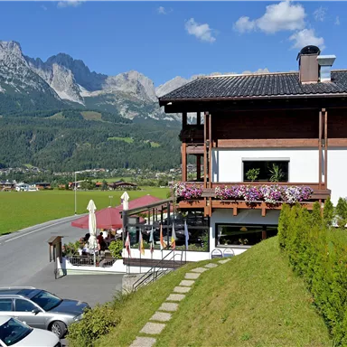 Ein malerisches Gasthaus mit Blumenbalkonen und einer herrlichen Sicht auf die Berge. Die Umgebung ist grün und einladend, ideal für einen gemütlichen Aufenthalt.