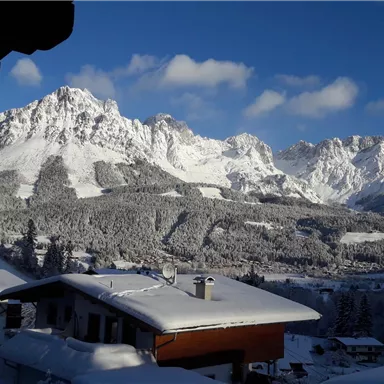 Eine verschneite Berglandschaft mit majestätischen Bergen im Hintergrund. Im Vordergrund sind schneebedeckte Dächer von Häusern zu sehen.