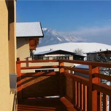 A balcony with a wooden railing that offers a picturesque view of snow-covered mountains. The sky is clear and blue, which impressively highlights the winter landscape.