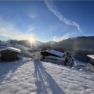 A snowy landscape with small wooden houses. The sun rises behind the mountains and the sky is clear.