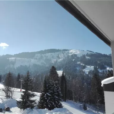 A winter landscape with snow-covered mountains and fir trees. The sky is clear and blue, creating a tranquil atmosphere.