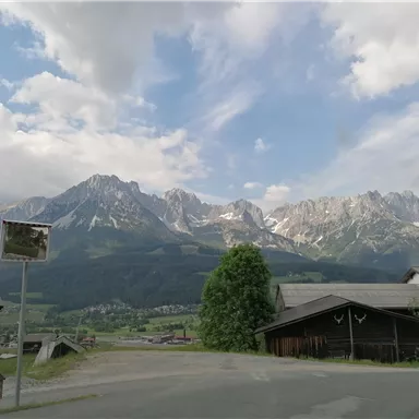 An impressive mountain landscape with high peaks and a blue sky. In the foreground, some buildings and trees can be seen.