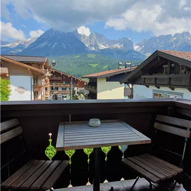 Ein gemütlicher Balkon mit einem Tisch und zwei Stühlen. Im Hintergrund sind beeindruckende Berge und eine malerische Landschaft zu sehen.