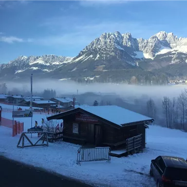 Eine verschneite Berglandschaft mit einem Holzhaus im Vordergrund. Im Hintergrund sind majestätische Berge und Nebel zu sehen.