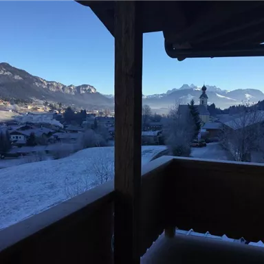 Eine winterliche Landschaft mit schneebedeckten Hügeln und Bergen im Hintergrund. Durch ein Fenster ist der Blick auf ein kleines Dorf und eine Kirche zu sehen.