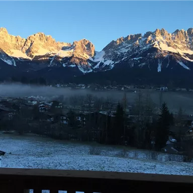Eine atemberaubende Berglandschaft mit schneebedeckten Gipfeln im Morgenlicht. Im Vordergrund ist ein bewaldetes Tal mit kleinen Häusern zu sehen.