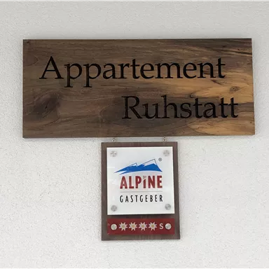 Ein Holzschild mit der Aufschrift „Appartement Ruhstatt“ und einem Logo von „Alpine Gastgeber“. Das Schild befindet sich an einer Wand und zeigt einen einladenden Unterkunftsbereich.
