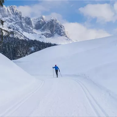 A cross-country skier on a snow-covered path in the mountains. The landscape is tranquil with snow-covered hills and a clear sky.