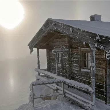 A snowy wooden cabin stands in a foggy landscape. The sun shines through the fog, creating a mystical atmosphere.