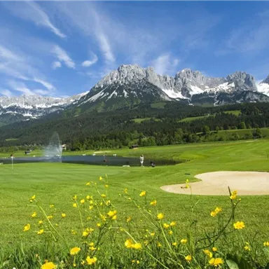 A picturesque golf course with green meadows and blooming flowers. In the background, majestic mountains and a lake with a water fountain can be seen.