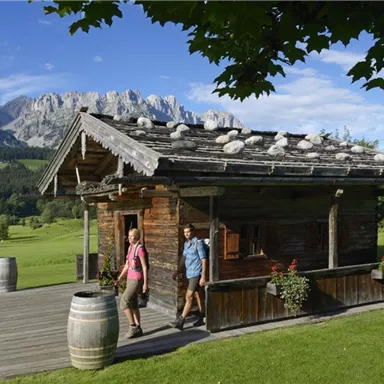A beautiful mountain cabin surrounded by green meadows and majestic mountains. Two people are leisurely walking to the cabin.