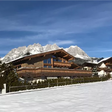 Eine malerische Winterlandschaft mit schneebedeckten Hügeln und traditionellen Chalets. Im Hintergrund sind beeindruckende Berge unter einem klaren blauen Himmel zu sehen.