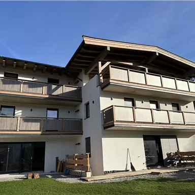 Ein modernes Gebäude mit mehreren Balkonen und einem Holz-Dach. Es steht in einer grünen Umgebung mit Bäumen im Hintergrund.