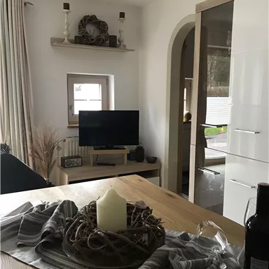 A cozy living space with a wooden table, a television, and a modern living wall. The decoration includes a candle and a stylish tablecloth.
