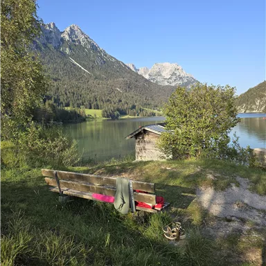 Ein ruhiger See umgeben von Bergen und Bäumen. Eine Bank lädt zum Verweilen und Genießen der Natur ein.