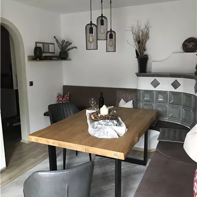 A cozy dining area with a wooden round table and modern chairs. In the background, decorative elements and a comfortable seating area can be seen.