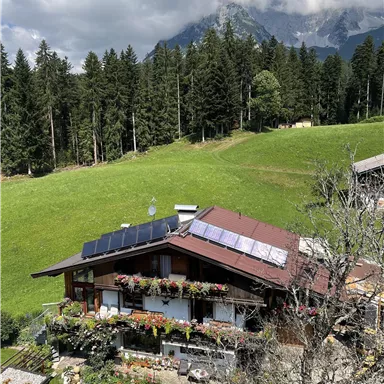 Ein charmantes Haus umgeben von grünen Wiesen und hohen Bäumen. Im Hintergrund ragen majestätische Berge unter einem teilweise bewölkten Himmel empor.