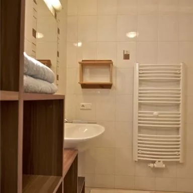 Ein modernes Badezimmer mit hellen Fliesen und einer Waschbecken. An der Wand befindet sich ein Handtuchwärmer und ein Regal mit gefalteten Handtüchern.