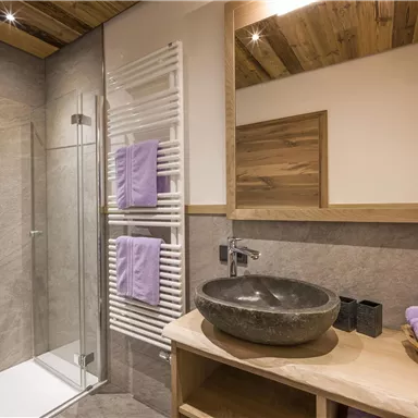 A modern bathroom with a glass shower and a stone sink. Towels in purple and wooden details give the space a cozy atmosphere.