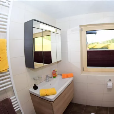 Ein modernes Badezimmer mit hellen Fliesen und einem großen Spiegel. Die bunten Handtücher und das Fenster sorgen für eine freundliche Atmosphäre.