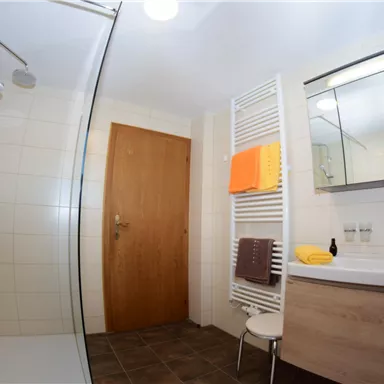 Ein modernes Badezimmer mit einer Dusche und einem Waschbecken. Die Wände sind hell gefliest und es gibt Handtücher in warmen Farben.