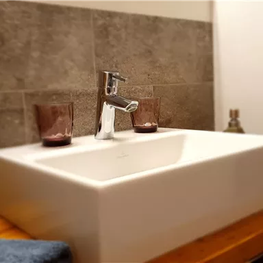 A modern sink on a wooden shelf. The wall is lined with gray tiles, and there are two decorative candles beside it.