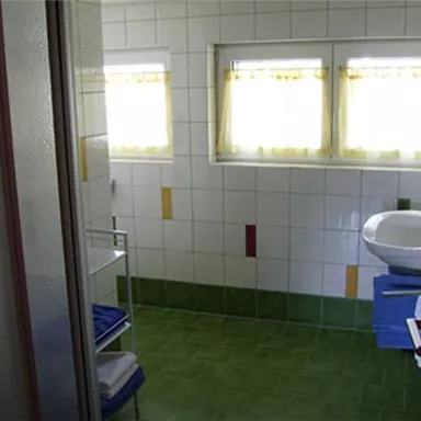 Ein helles Badezimmer mit weißen Fliesen und bunten Akzenten. Es gibt ein Waschbecken, Handtücher und große Fenster mit gelben Vorhängen.